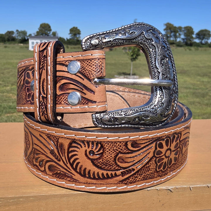 Brown leather belt with intricate designs and a silver buckle on a wooden surface with a grassy field and trees in the background.